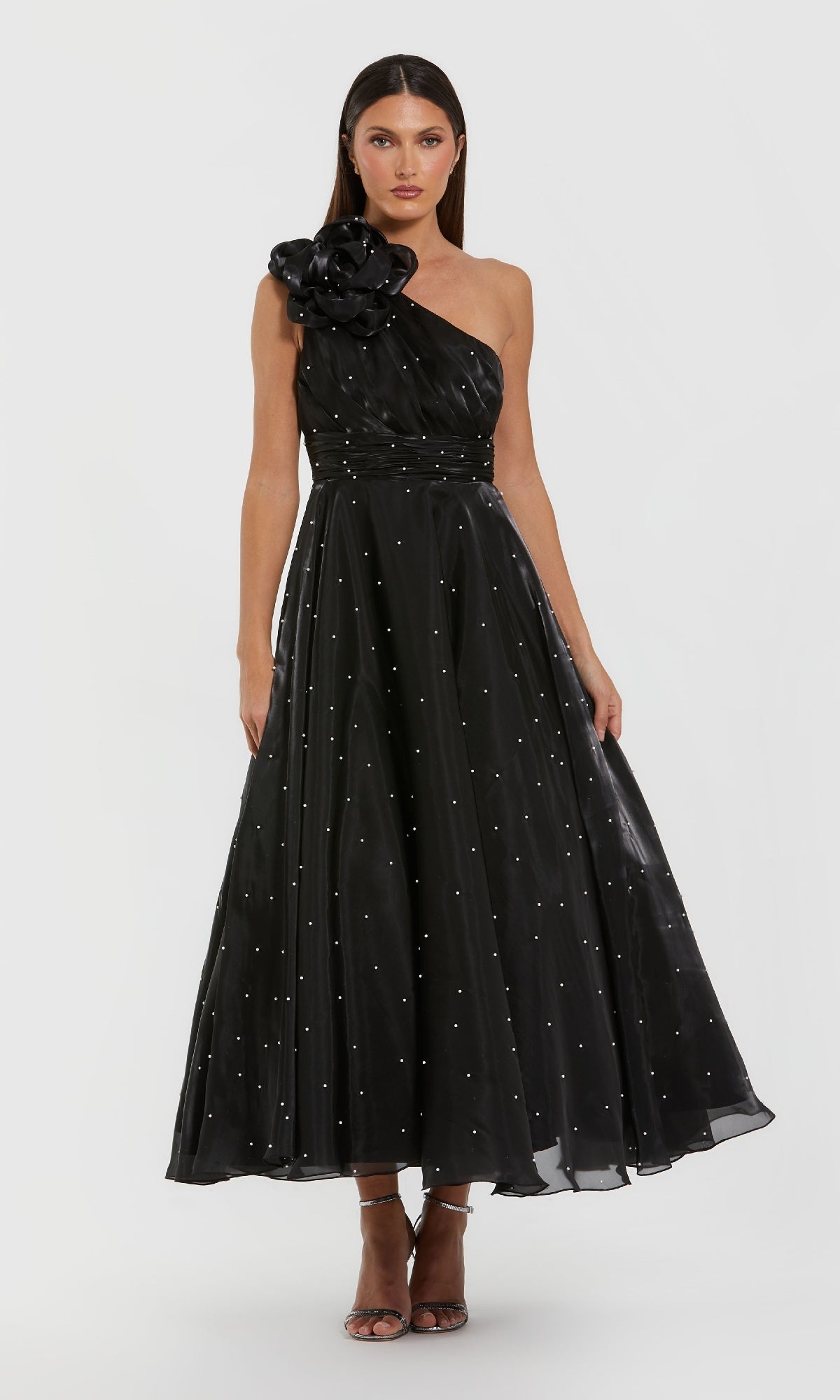 A woman wears the Mac Duggal 50078 one-shoulder black formal dress featuring pearl beading, a large floral shoulder detail, scattered embellishments, and pairs it with open-toe heels.